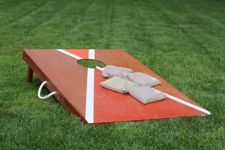 Corn hole board sitting on grass with four light purple bean bags sitting on top
