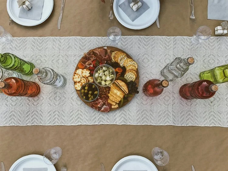 Charcuterie board onto of while patterned table runner with clear and red glass bottles around it