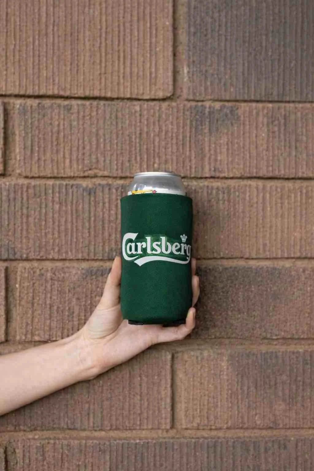Carlsberg koozie holding a drink inside and a hand holding the koozie on a brown wall background