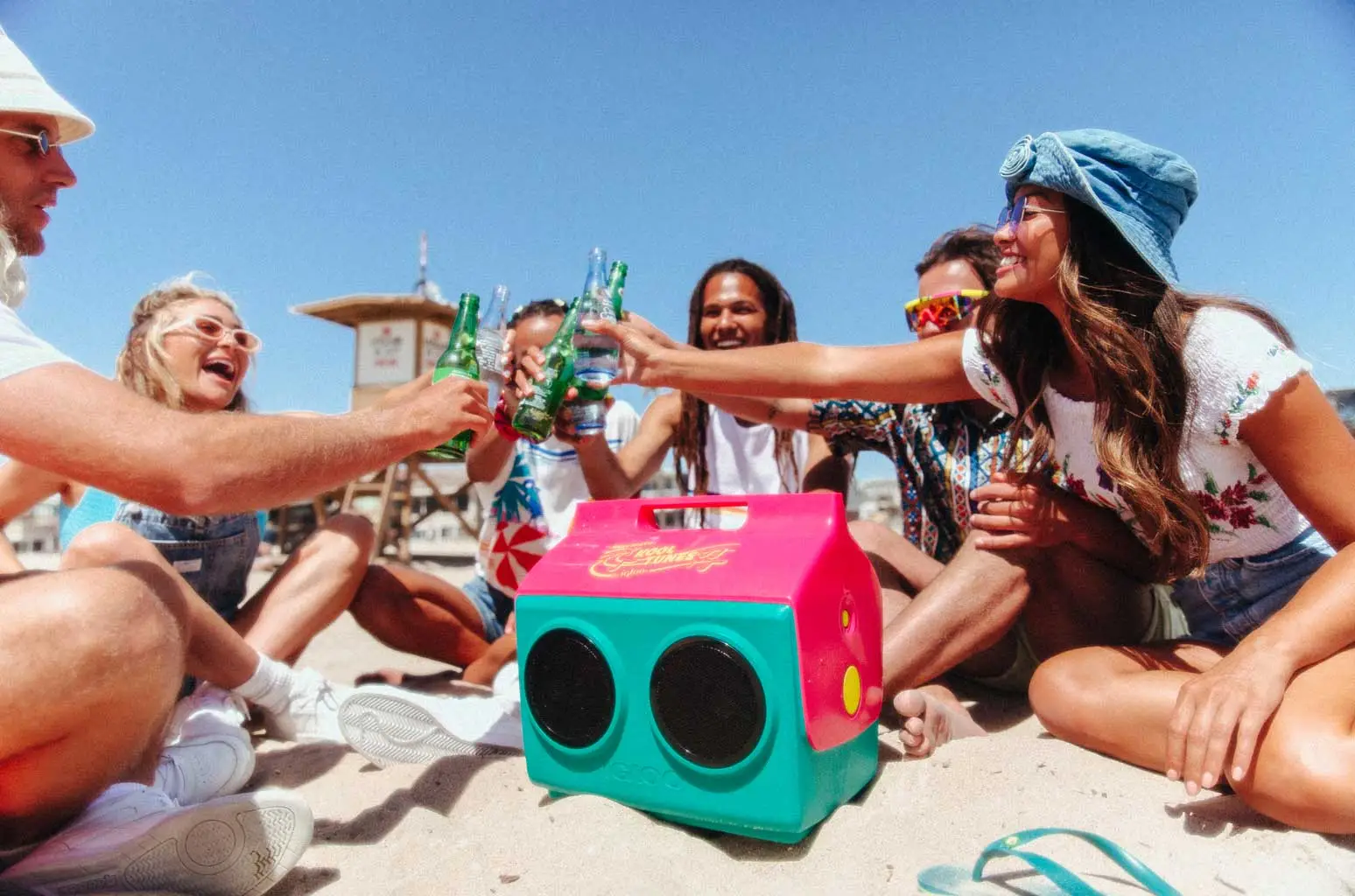 A pink and green Cooler Box with Speakers is places around people having fun and drinking on a a beach