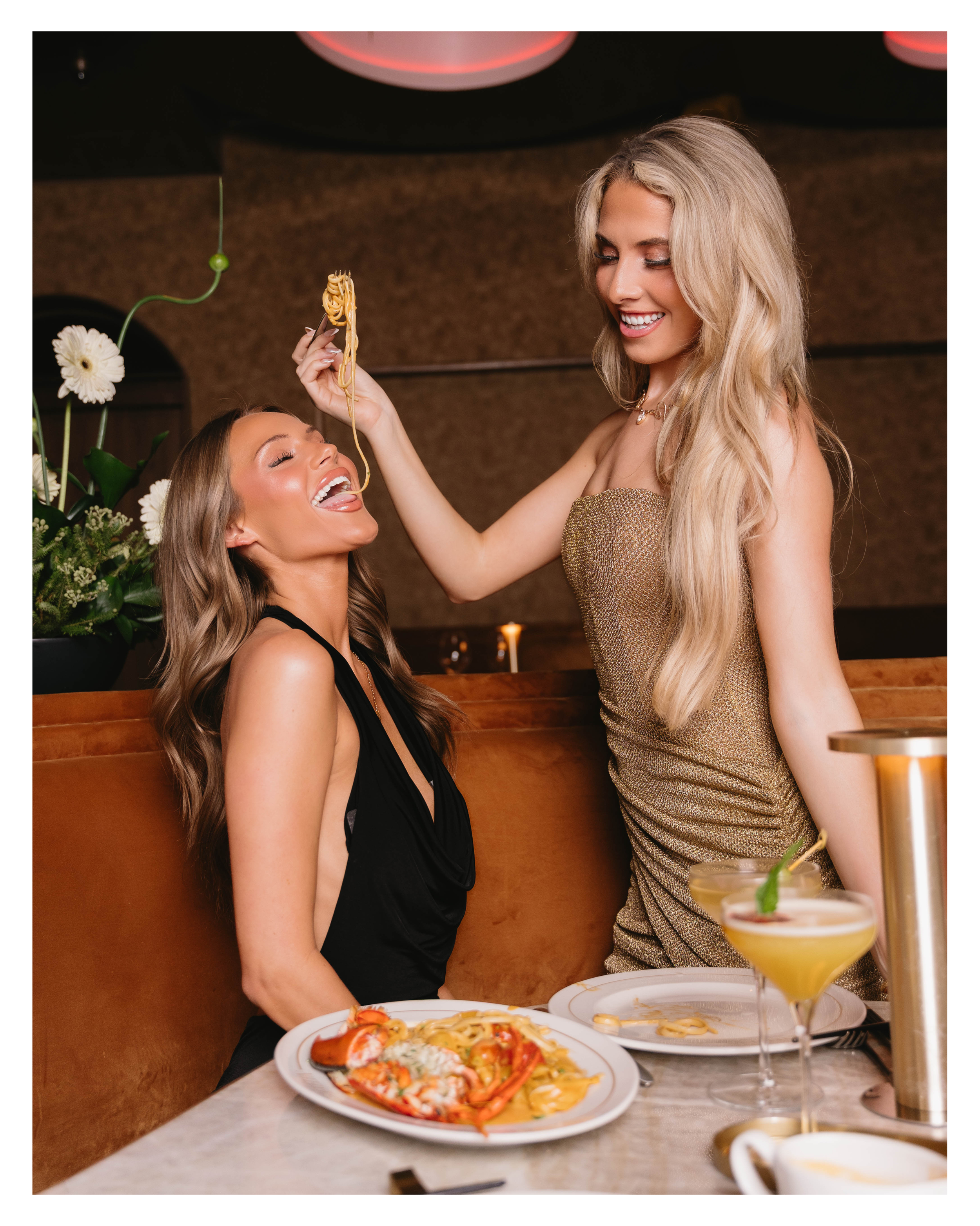 Two friends laughing in a velvet booth sharing lobster pasta during a girls night out at EVELYN Dallas