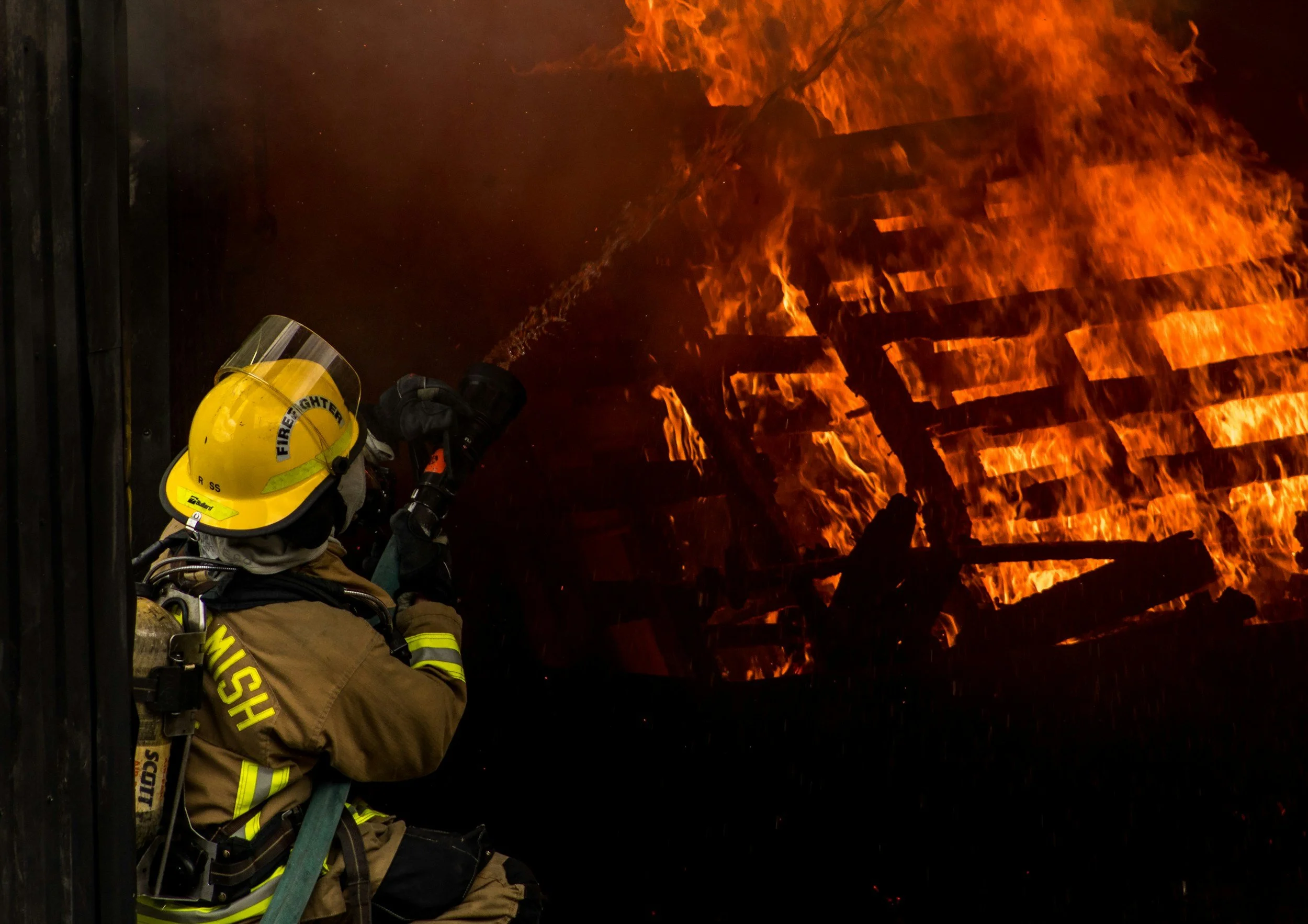Firefighter in protective gear, including a yellow helmet and tan uniform, uses a hose to fight a large, intense forest fire with bright orange flames and thick smoke at night.