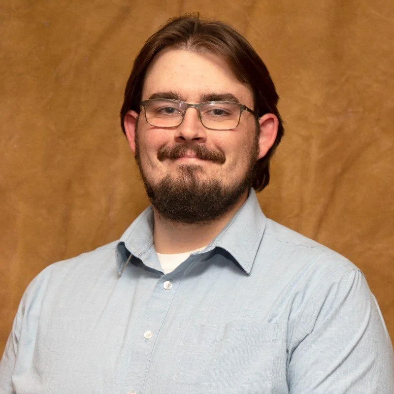 A man with glasses, dark hair, and a beard, wearing a light blue collared shirt, standing in front of a brown textured background.
