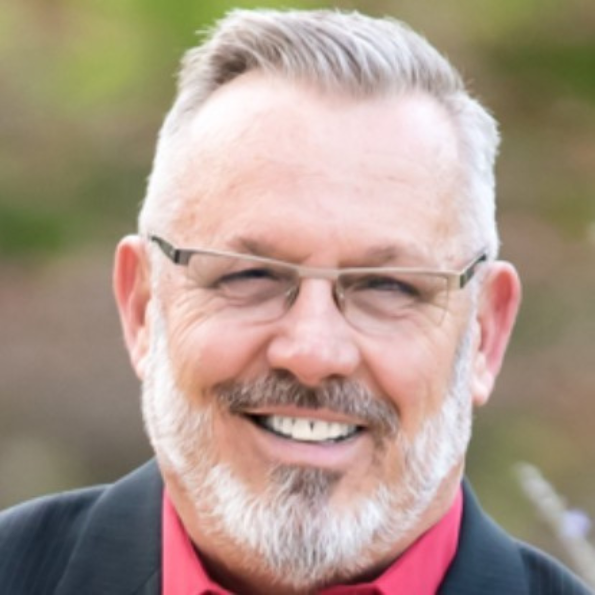 Portrait of a smiling man with glasses, gray hair, and a beard, wearing a dark suit and red shirt, outdoors.