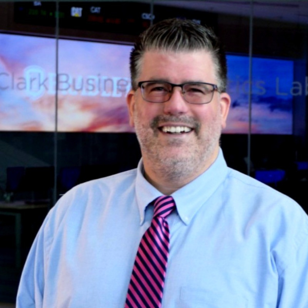 A smiling man wearing glasses, a light blue shirt, and a pink striped tie, standing in front of a digital screen with the text 'Clark Business' and a cloudy sky background.