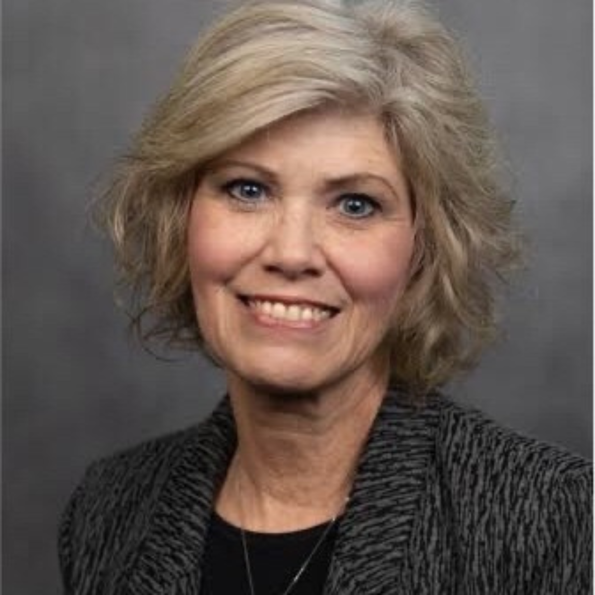 A headshot of a smiling middle-aged woman with blonde, wavy hair and blue eyes, wearing a dark top and a black and white striped blazer.