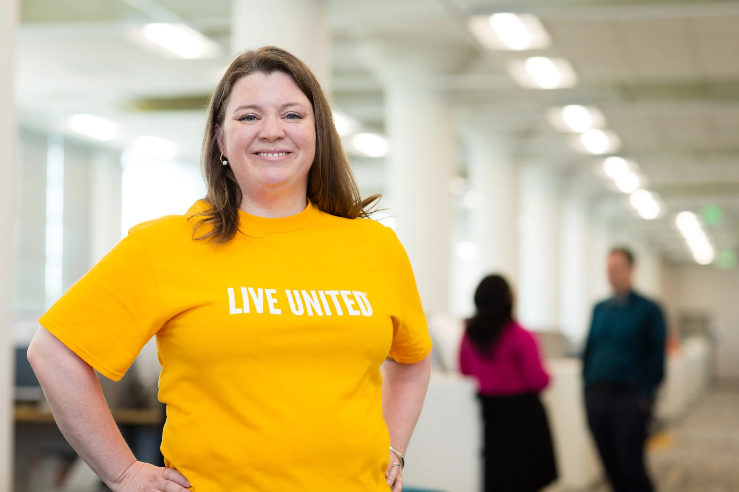 A woman with shoulder-length brown hair smiling, wearing a yellow T-shirt with 'LIVE UNITED' written on it, standing in an open indoor office or community space with blurred people in the background.