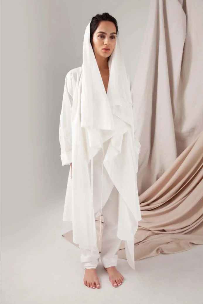 A person wearing a long, flowing white outfit with a hood, standing barefoot against a backdrop of draped fabric.
