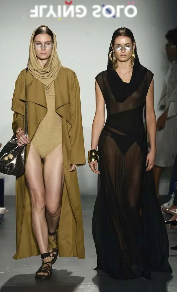 Models walking on a fashion runway wearing layered clothing, including a brown hooded cape and a sheer black dress, accessorized with gold jewelry.