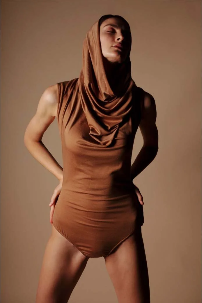 Person wearing a brown bodysuit with a draped hood, posing with hands on hips against a plain background.