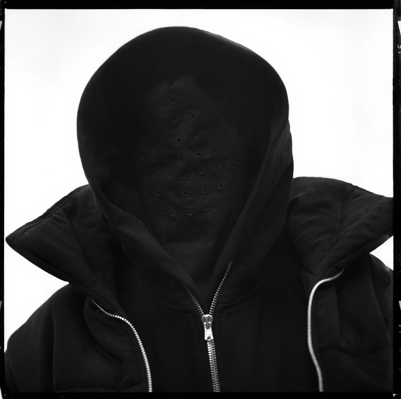 Person wearing a black hoodie with a face covering featuring holes, creating a mysterious appearance against a plain background.