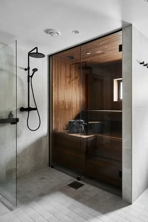 Tummaksi paneloitu pieni sauna, jossa on lasiseinä, ja vaalea kylpyhuone mustalla suihkusetillä.