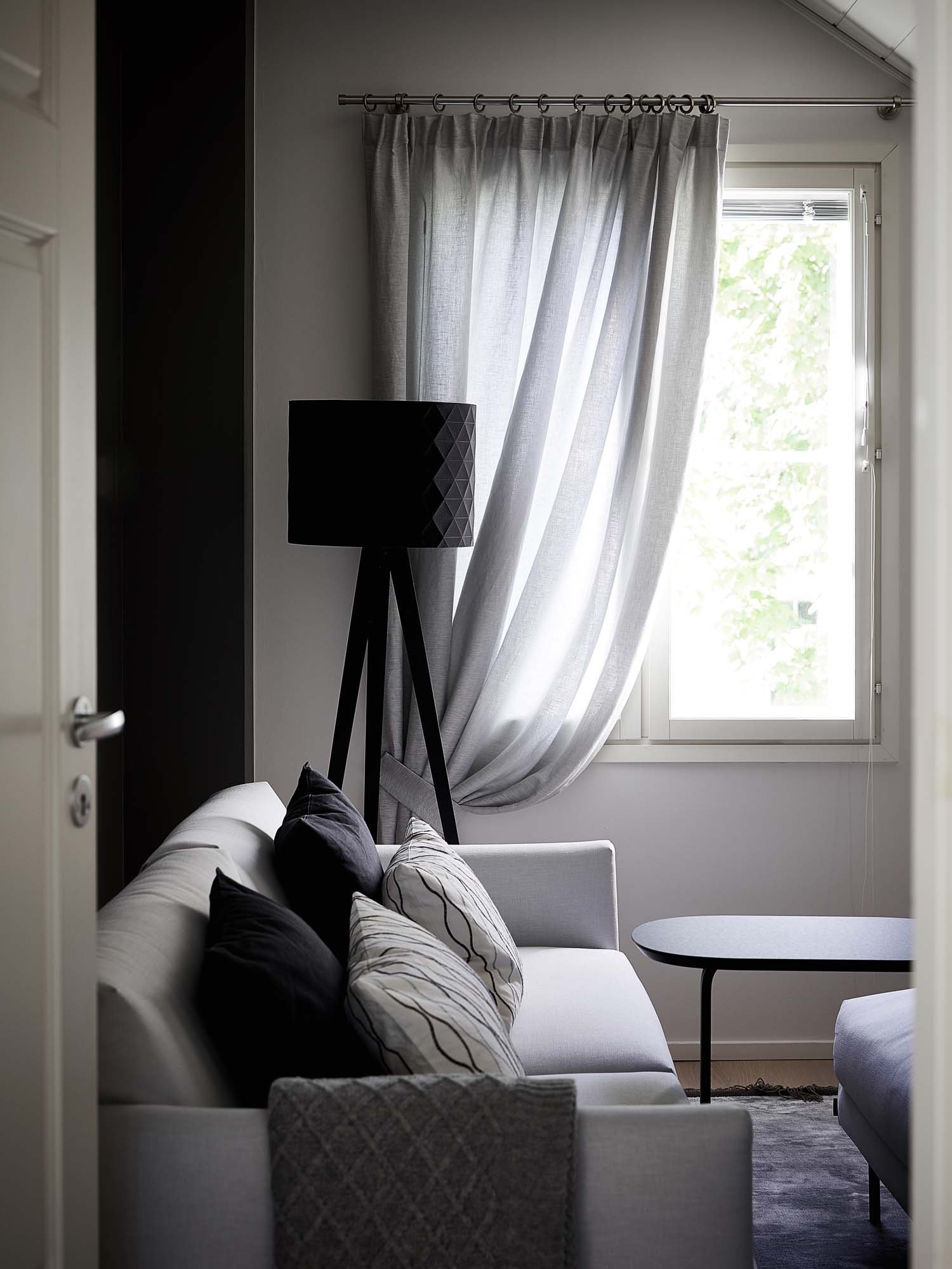 Moderni olohuone, jossa harmaa sohva, koristetyynyjä, musta jalkalamppu, vaaleat verhot ja ikkuna, josta näkyy vehreää luontoa. Pöydällä on pöytävalo ja sisustus on vähäeleinen.