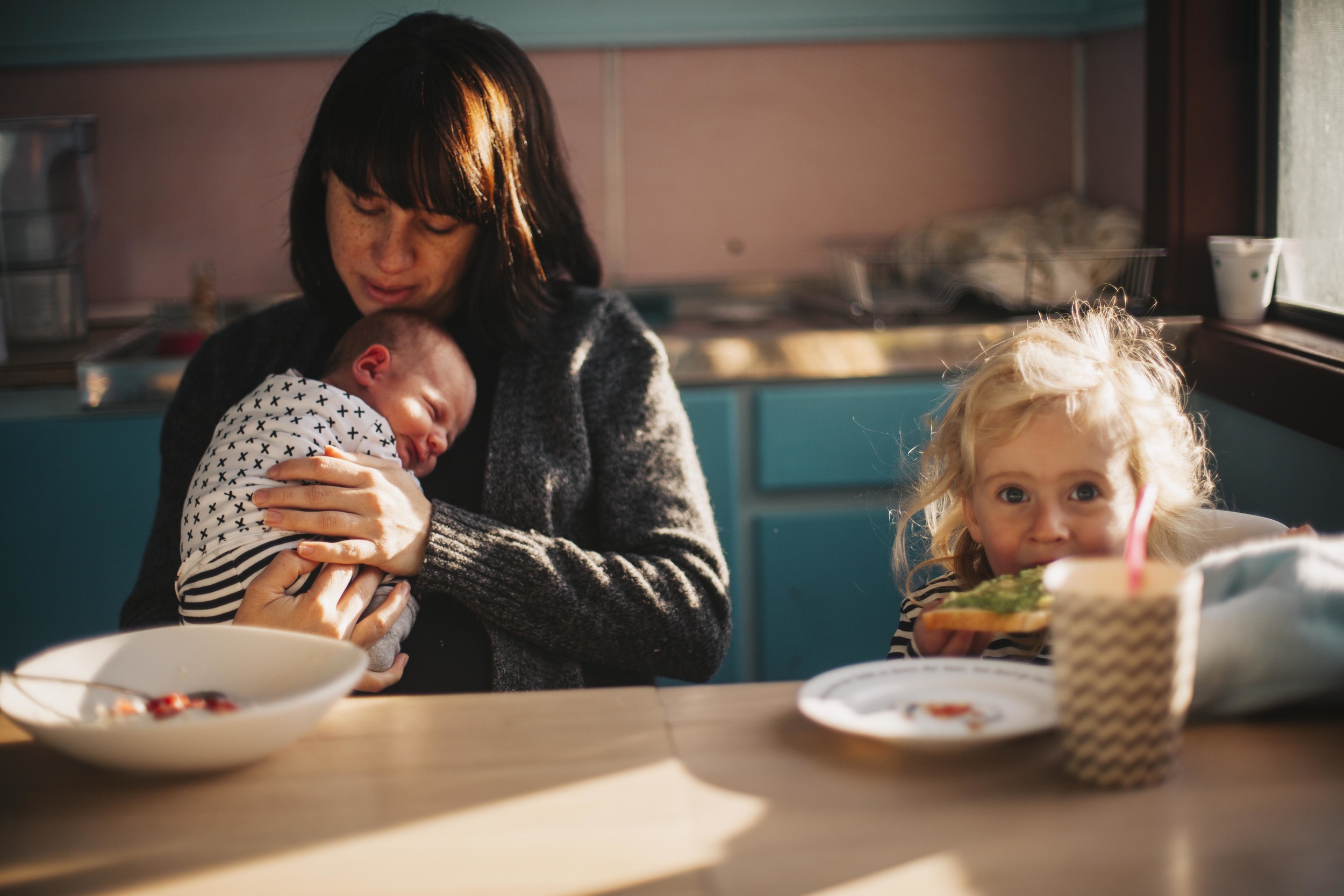 mother holding newborn baby while daughter eats toast at kitchen table