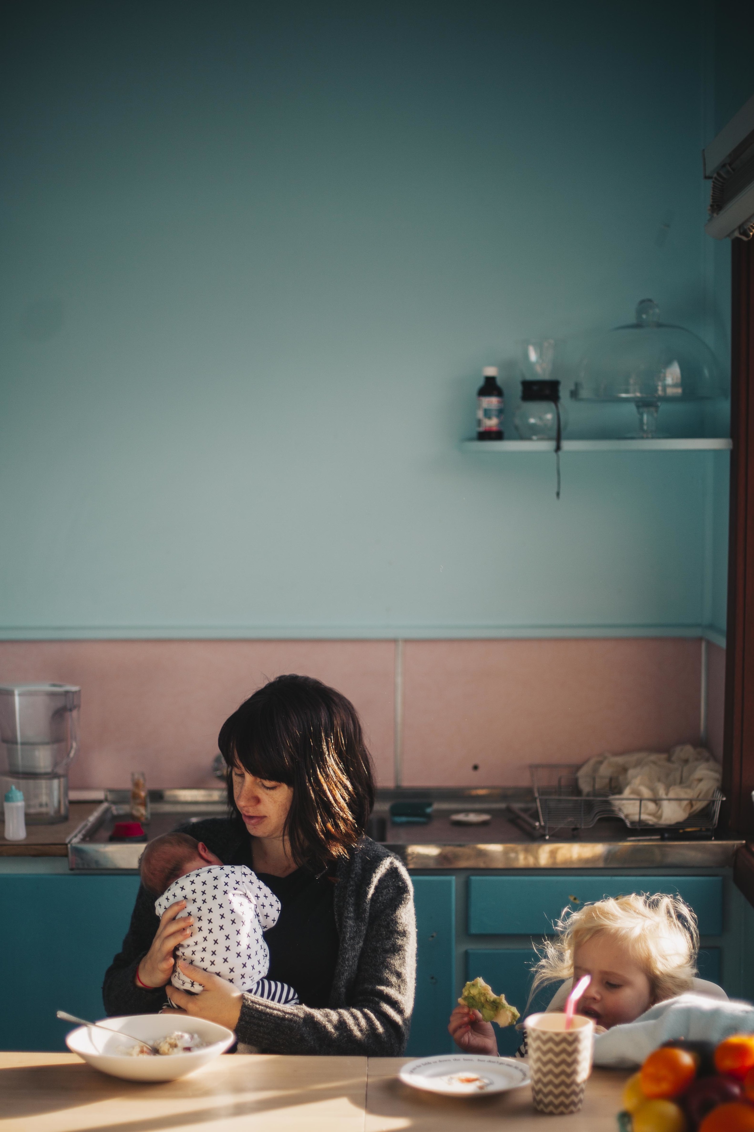 mother holding newborn baby while daughter eats toast at kitchen table
