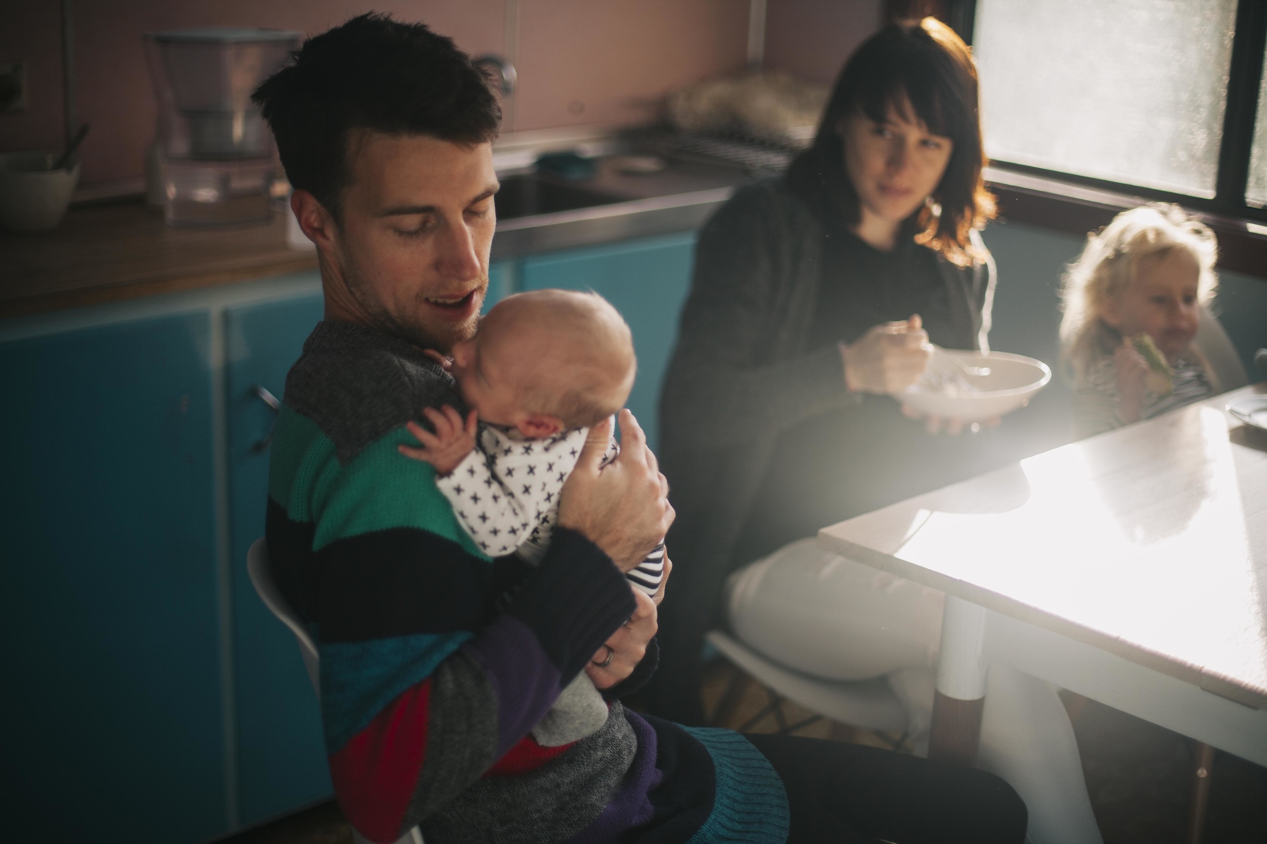father holding newborn baby while daughter eats toast at kitchen table
