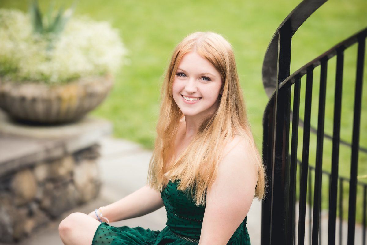 A smiling high school senior with long red hair sitting outdoors near a black metal railing and a stone planter in the background.