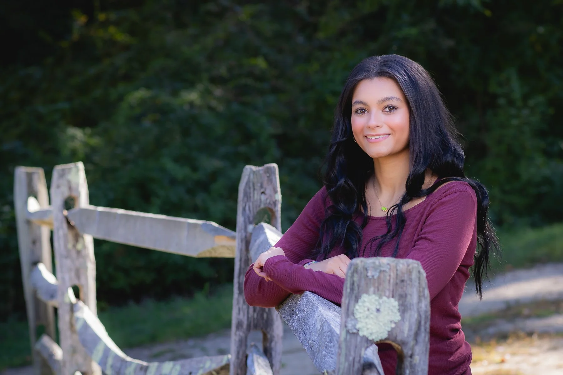 A woman with long black hair, wearing a maroon long-sleeved shirt, leaning on a rustic wooden fence outdoors, smiling, with a background of green trees.