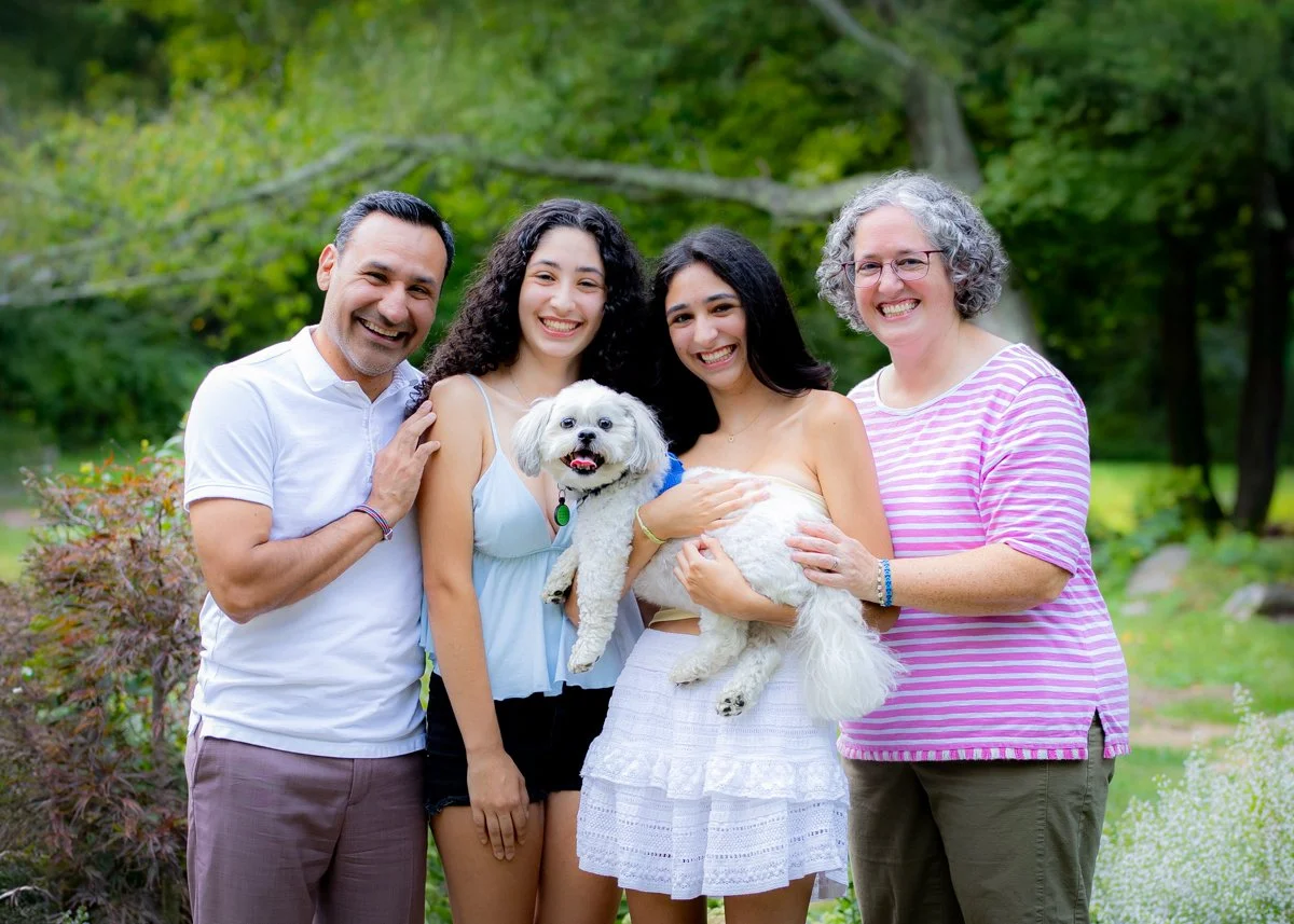 Group of five people smiling outdoors, two women holding small white dog, in a lush green park.