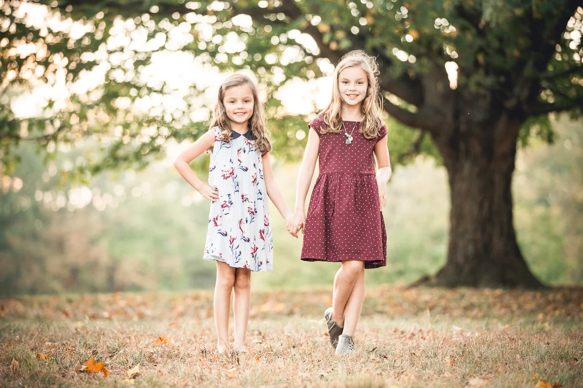 Two young girls holding hands and smiling in a park with a large tree in the background during fall.