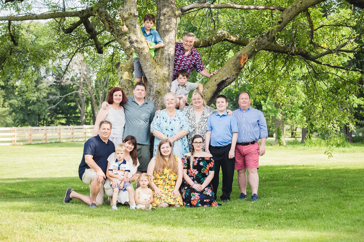 A large family of 14 people gathered outdoors in front of a big tree with green leaves, smiling and posing for a photo on a sunny day.