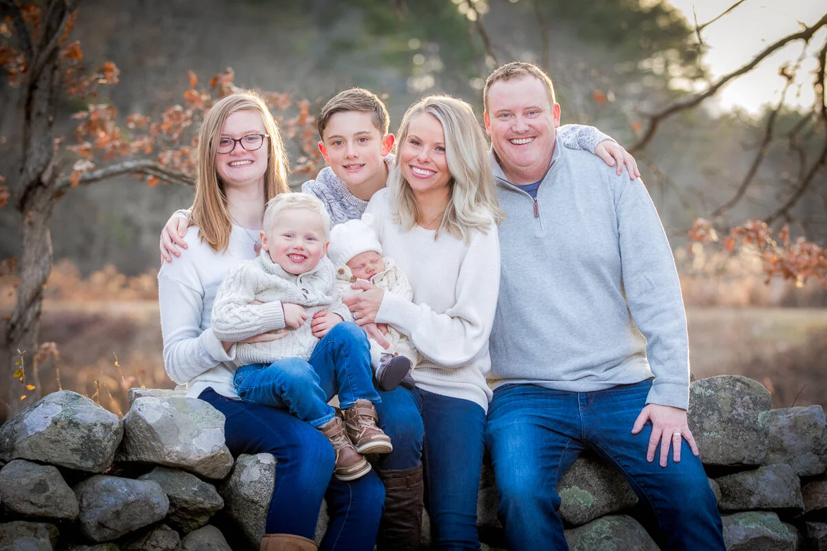 A family of six sitting on a stone wall at the Grist Mill in Sudbury, smiling and posing for the photo during fall.