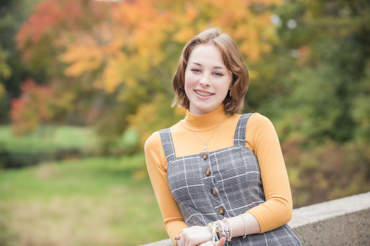 A young woman with short light brown hair and braces standing outdoors with colorful autumn trees in the background, smiling at the camera. She is wearing a yellow turtleneck sweater layered under a gray checkered dress with buttons and has jewelry o