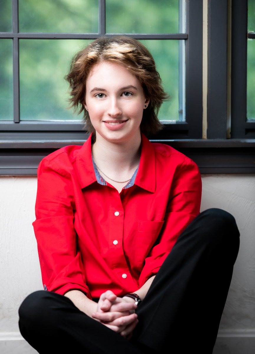 a high school senior with short brown hair and a red shirt sitting by a window and smiling.