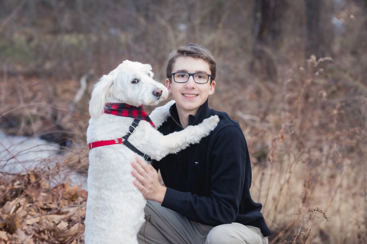 A young man wearing glasses and a black jacket sitting outdoors with a white dog that has a red and black checkered bandana. The dog is standing on its hind legs with its front paws on the man's shoulders, and the background shows dry brown leaves an