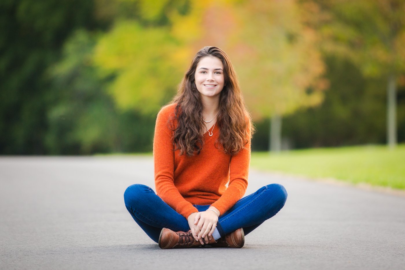A high school senior with long curly brown hair sitting cross-legged on an empty paved road outdoors, smiling, wearing an orange sweater, blue jeans, and brown shoes, with a background of green and autumn-colored trees.