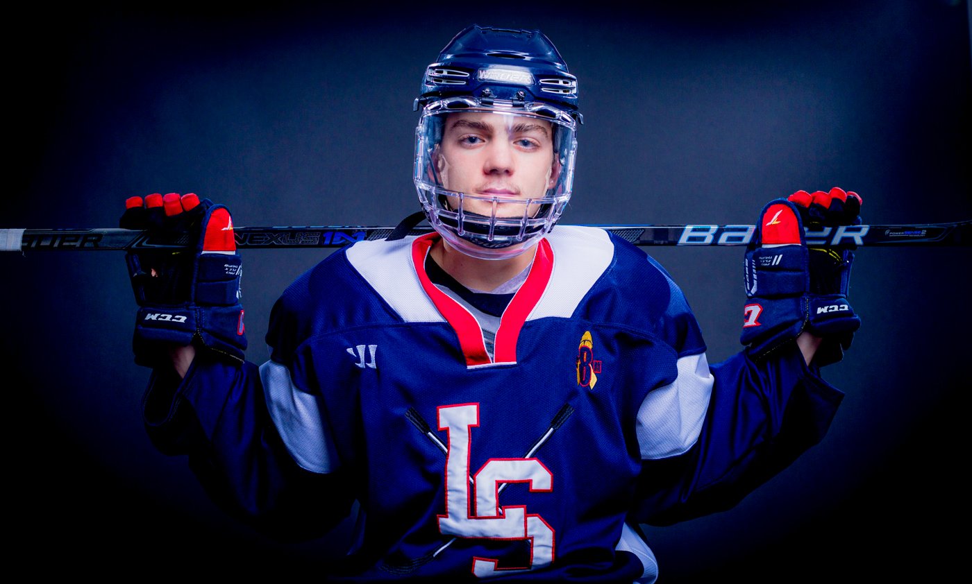 A High School Senior hockey player in helmet and jersey holding a hockey stick over his shoulders, facing forward.