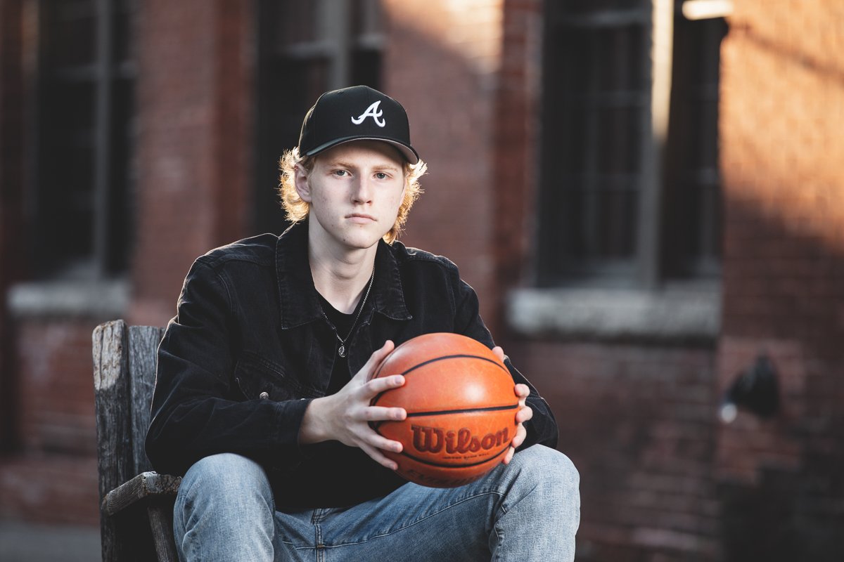 high school senior sitting outdoors holding a Wilson basketball, wearing a black baseball cap and black jacket, with a focused expression.