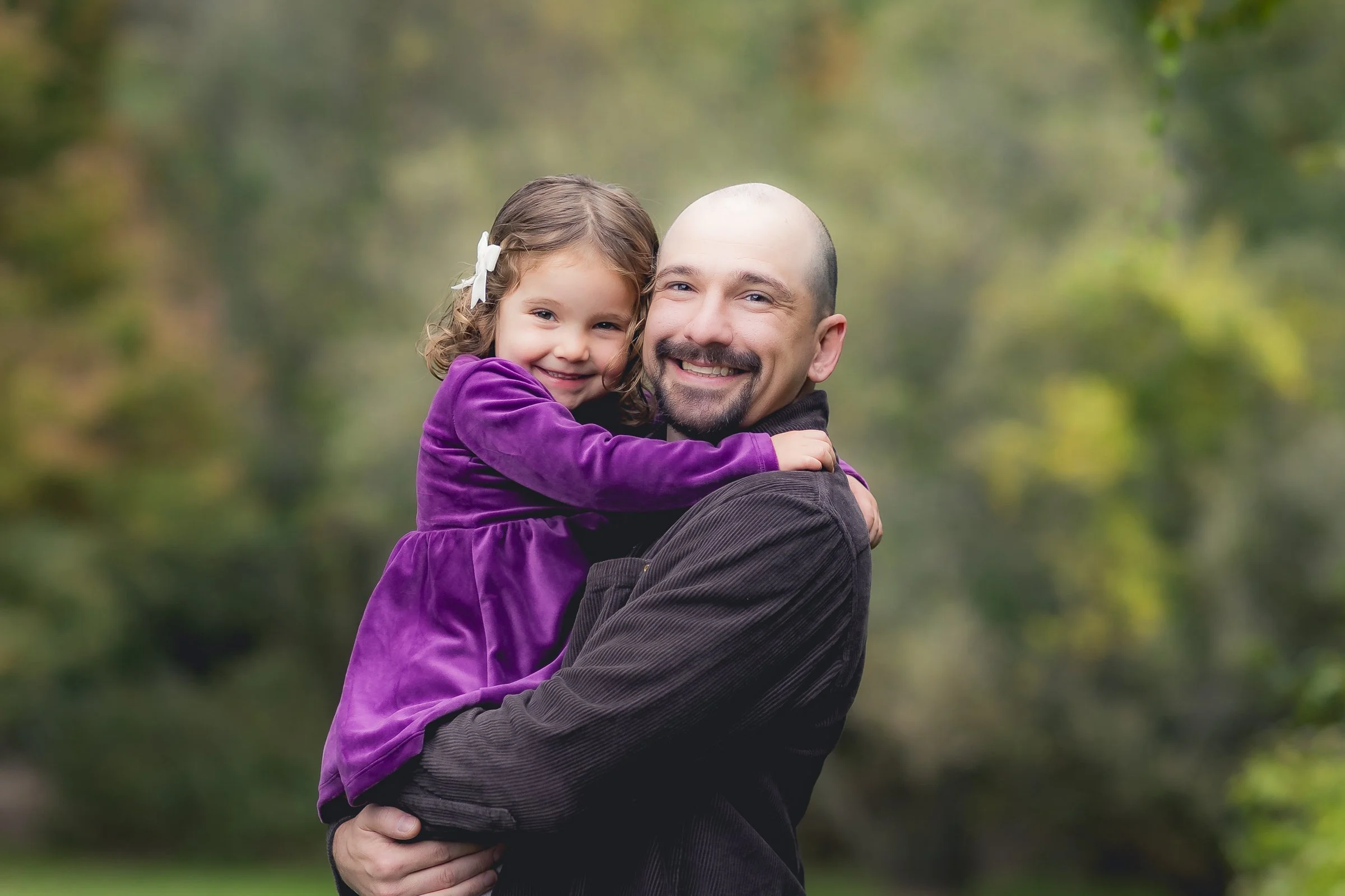A father holding his daughter in an outdoor park in Sudbury, MA, with green trees in the background. The girl is smiling, wearing a purple dress, and has a white bow in her hair. They are hugging and smiling at the camera.