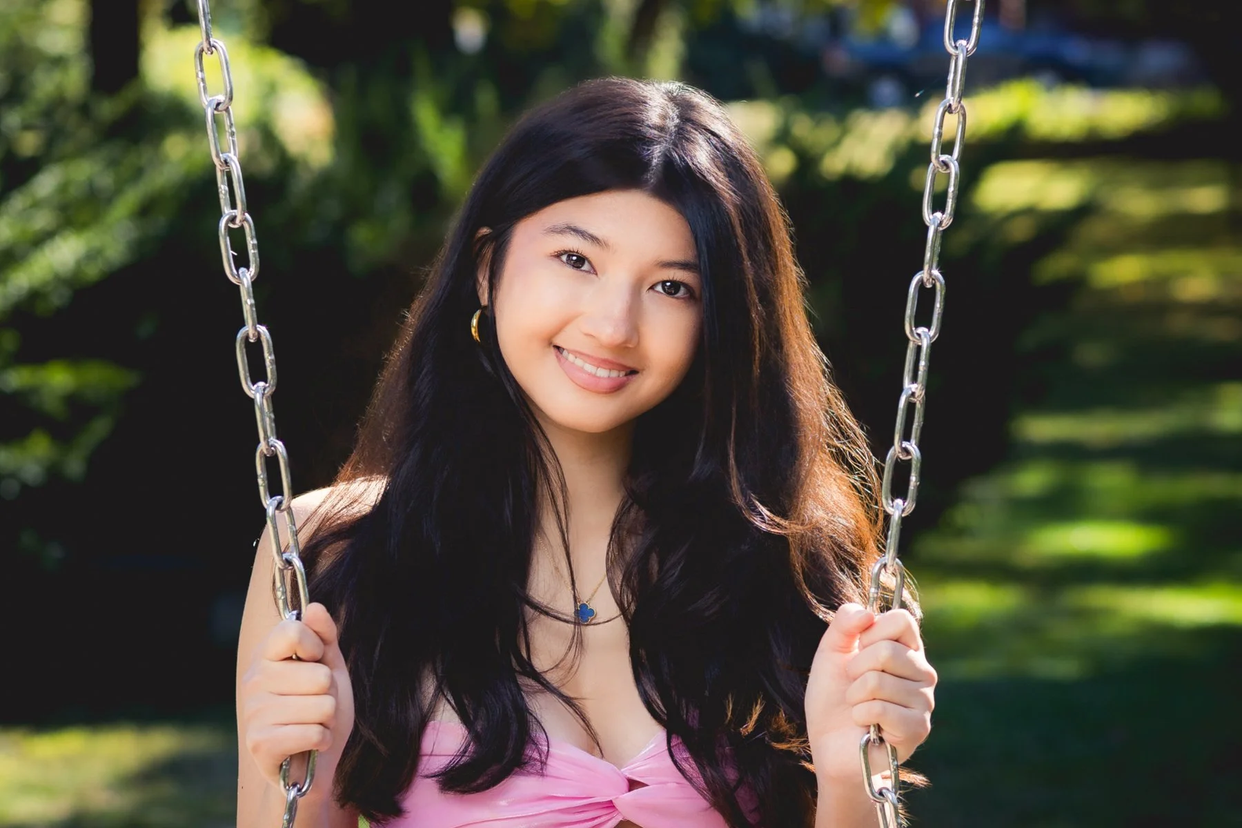 A high school senior in Sudbury Grist Mill with long black hair smiling while holding onto a metal chain on a swing outdoors.