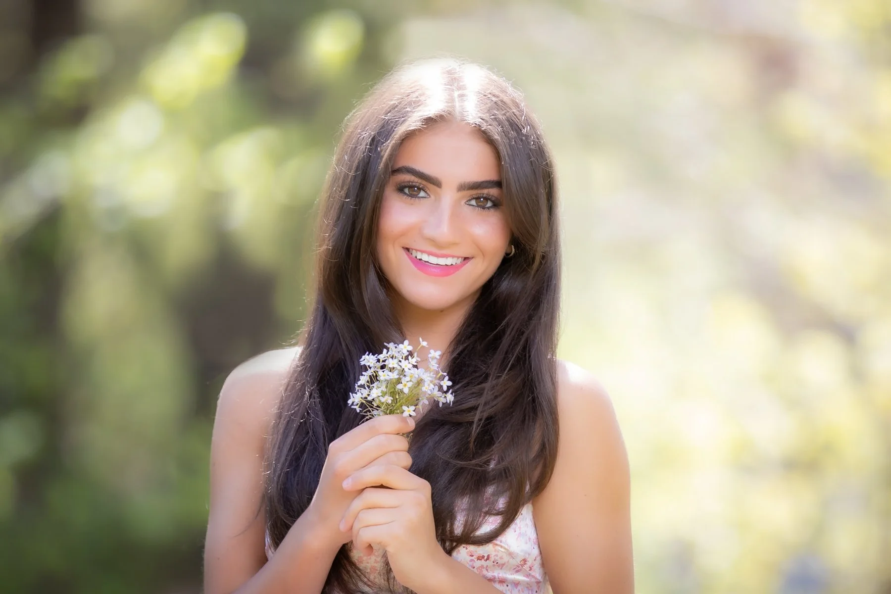 Smiling high school senior from Lincoln Sudbury Regional High School with long dark hair holding a small bouquet of white flowers outdoors on a sunny day.