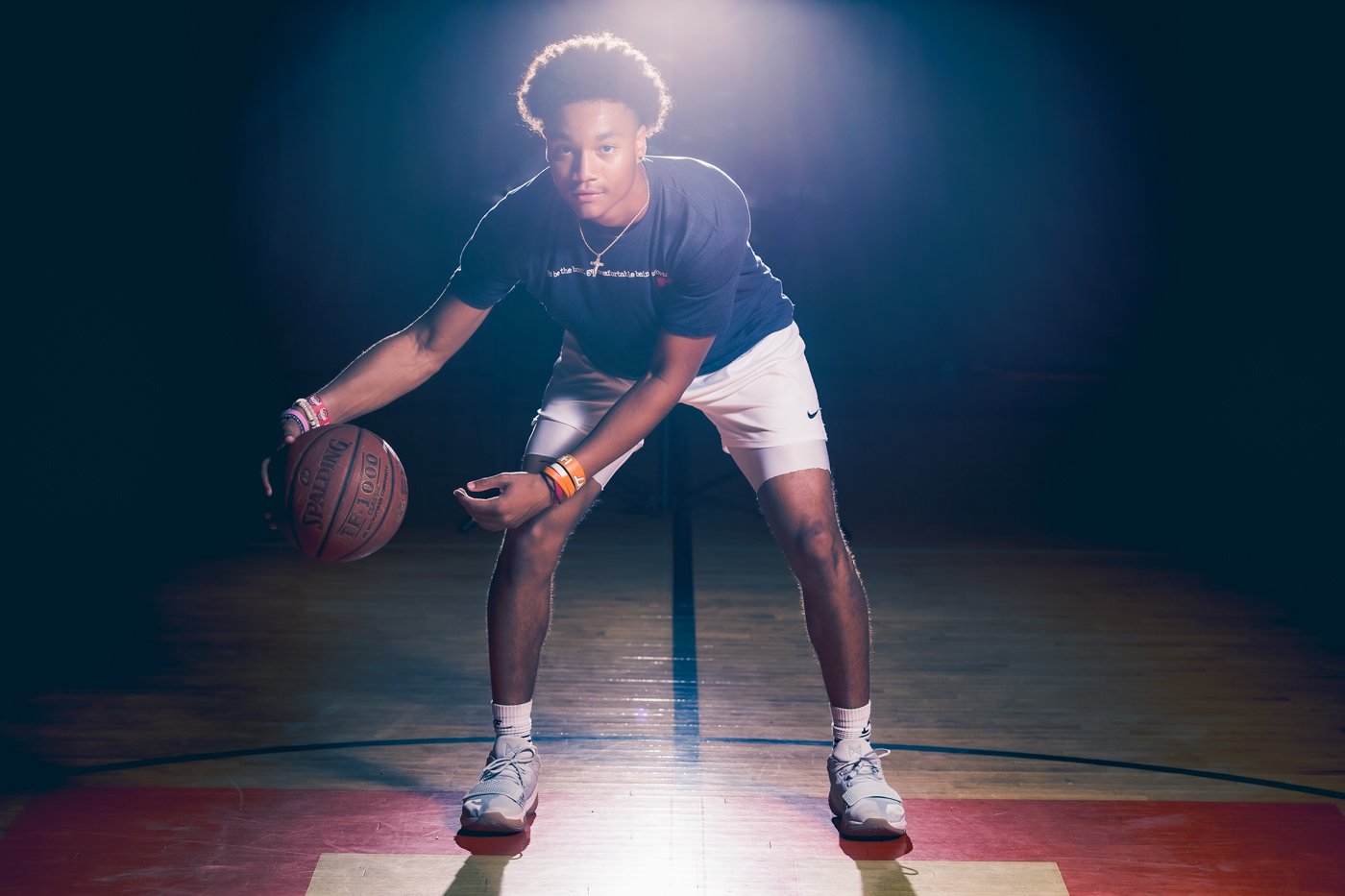 A high school senior basketball player in a dark gymnasium prepares to dribble a basketball on a lit court.