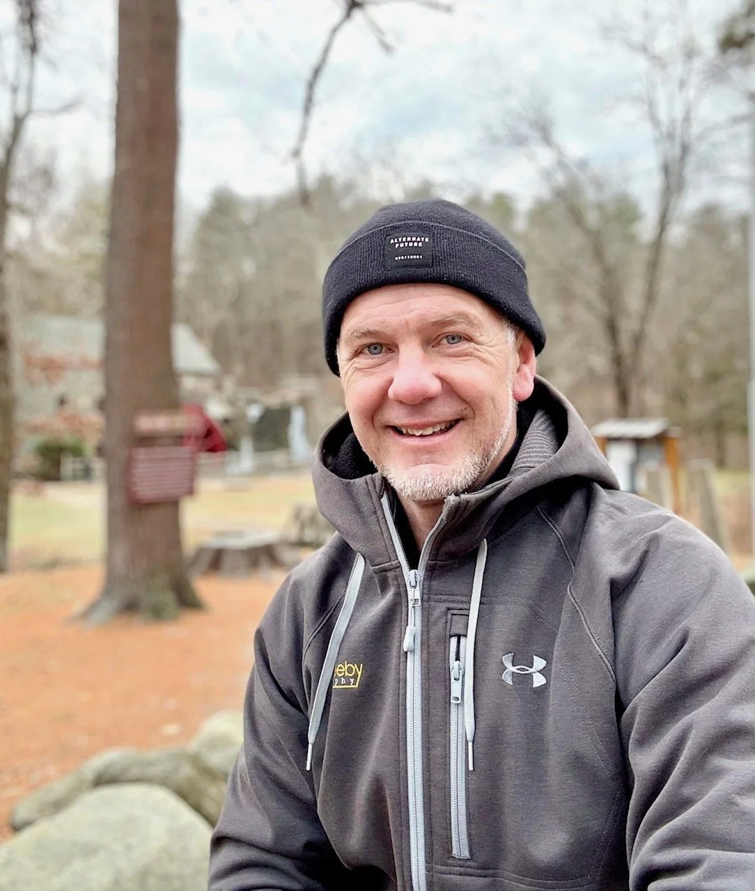 Stuart Beeby outdoors wearing a black beanie and gray jacket, smiling at the camera. Background shows trees and a park.