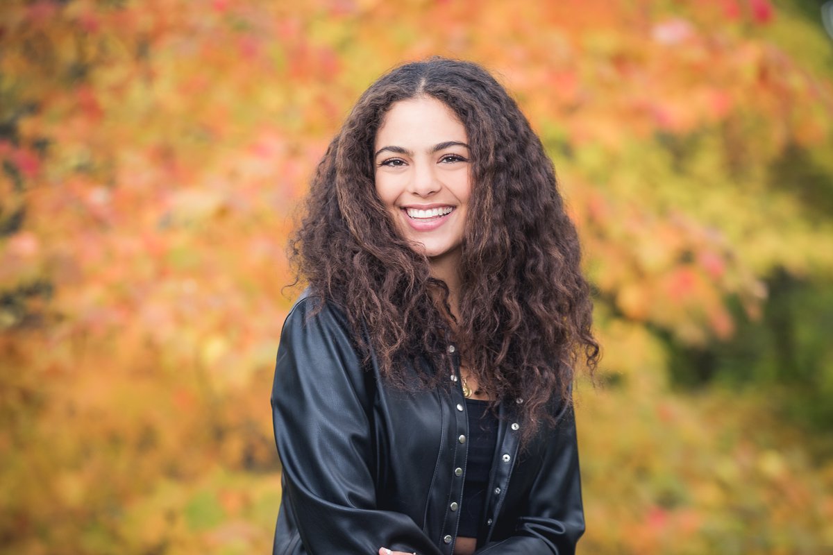 A high school senior with curly brown hair smiling outdoors with fall foliage in the background.