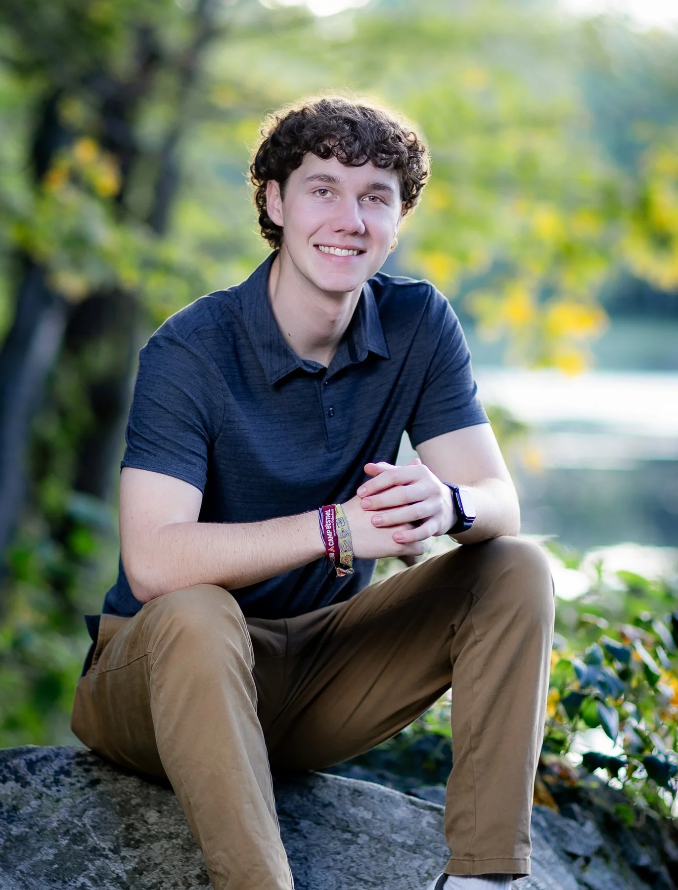 A high school senior with curly brown hair, wearing a dark blue polo shirt and tan pants, sits on a rock outdoors near a body of water surrounded by trees, smiling at the camera.