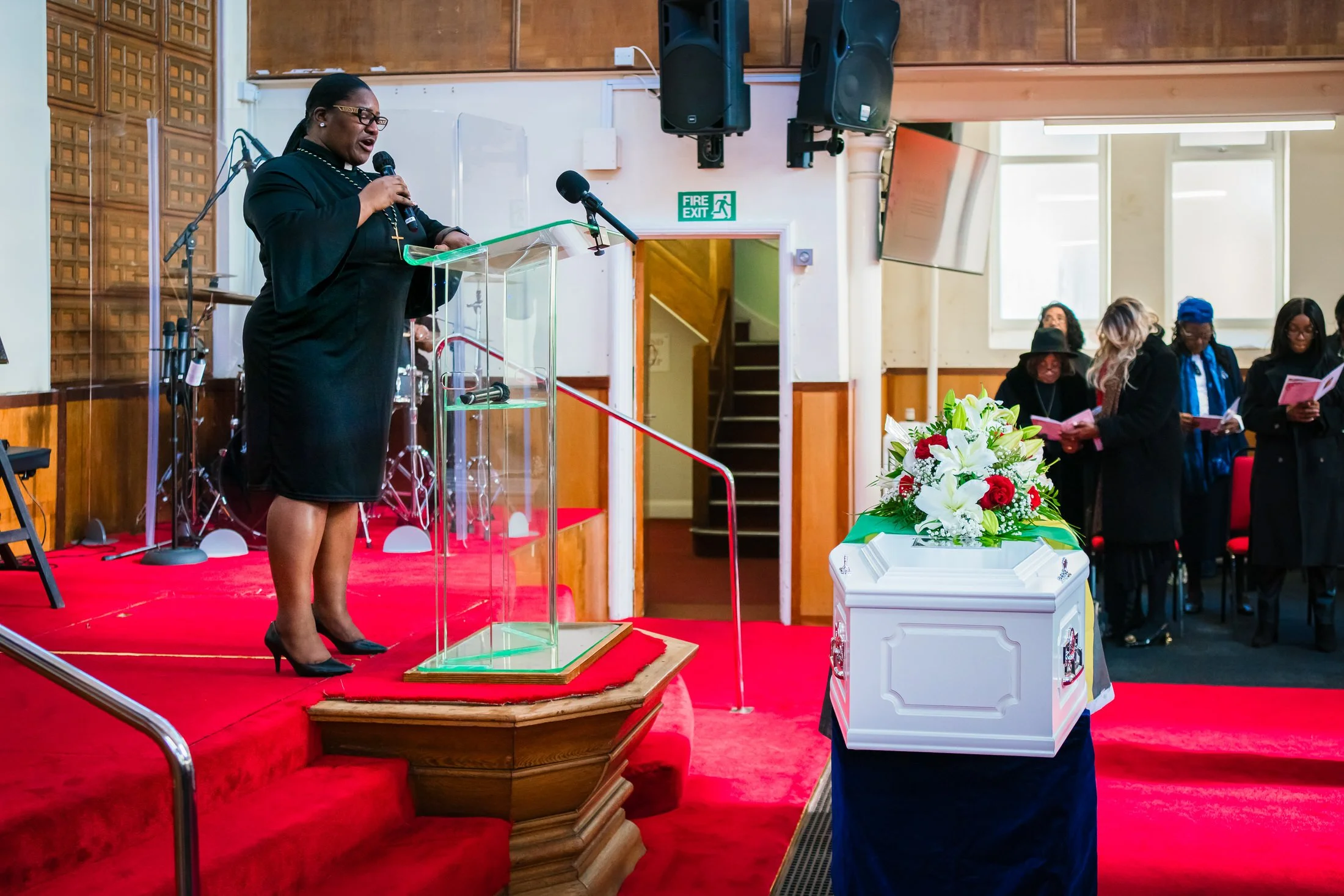 Woman in black speaking at a clear lectern beside a white coffin topped with white lilies and red roses, with mourners holding orders of service
