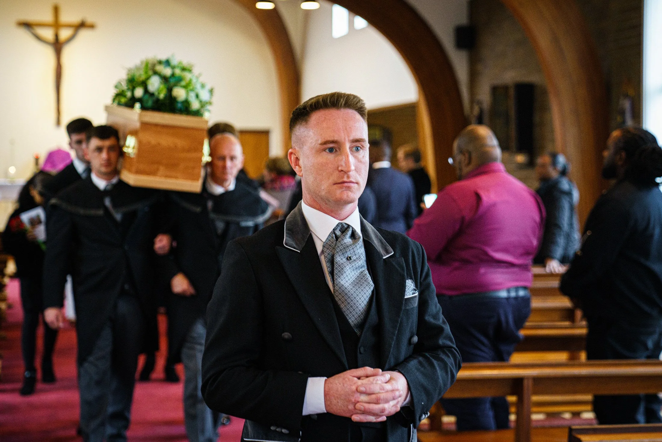 Bearers carrying a coffin with white roses out of church behind the funeral director, photographed by funeral photographer Shaun Foulds