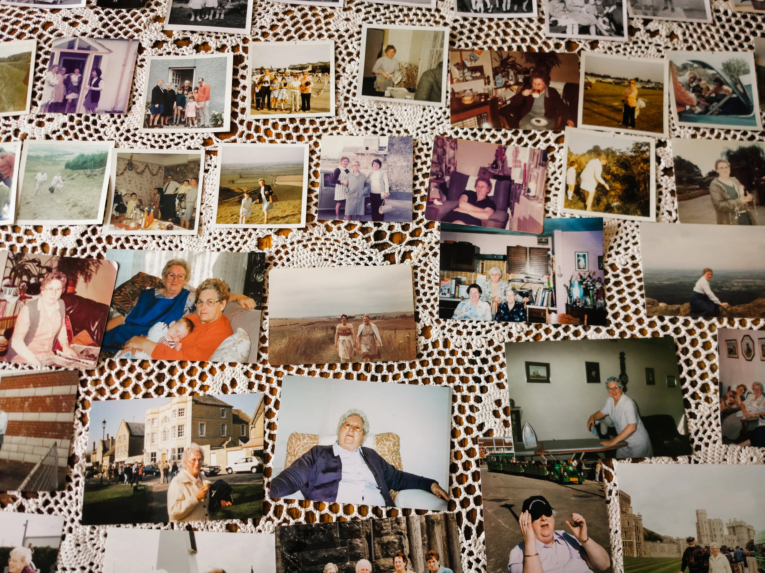 Dozens of family photographs spanning a lifetime, from holidays to living rooms, laid out on a white lace tablecloth, ready to be made into a funeral slideshow.