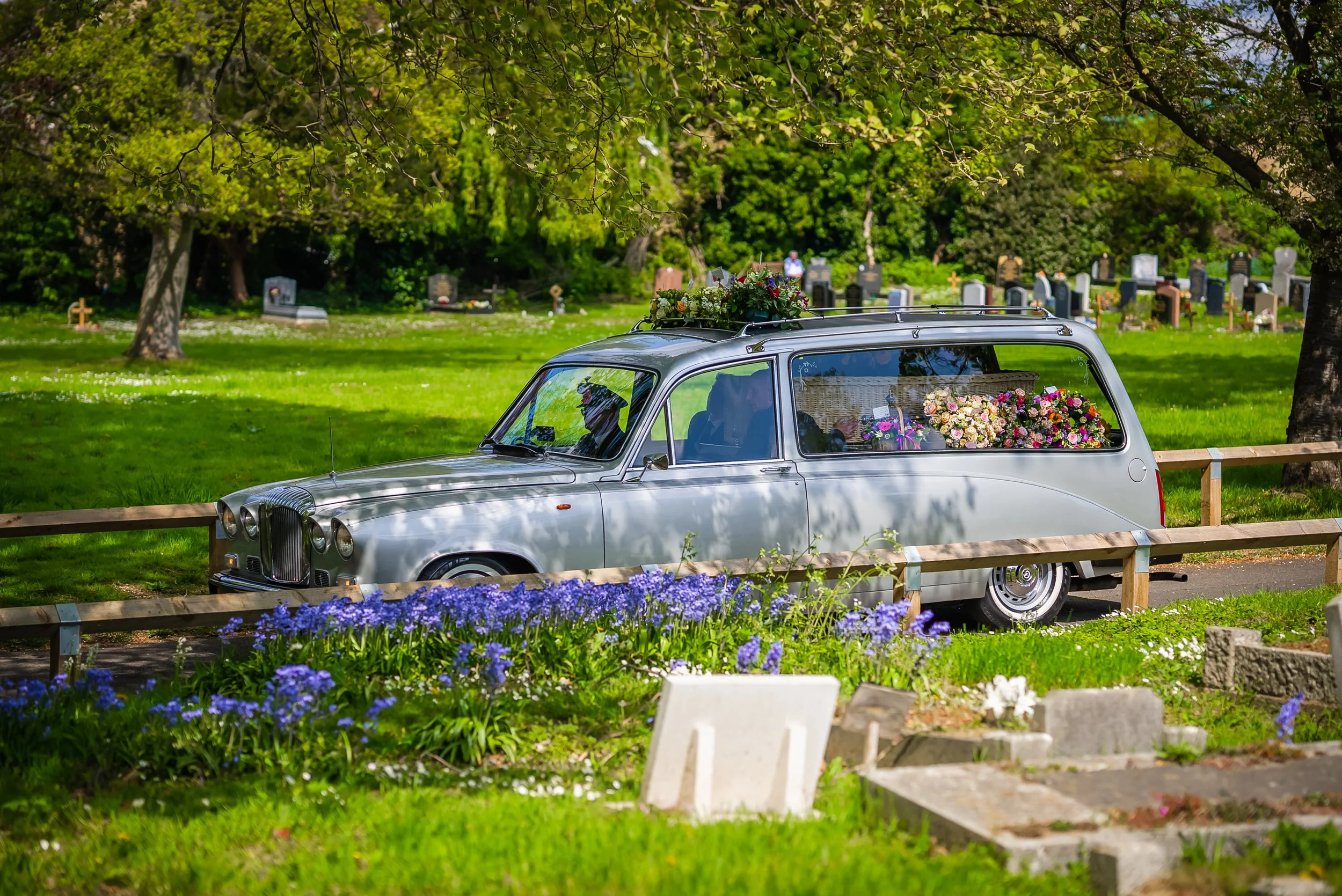 A silver hearse carries a flower-covered coffin through Richmond Cemetery in London, past spring bluebells, on the way to a funeral I live streamed.