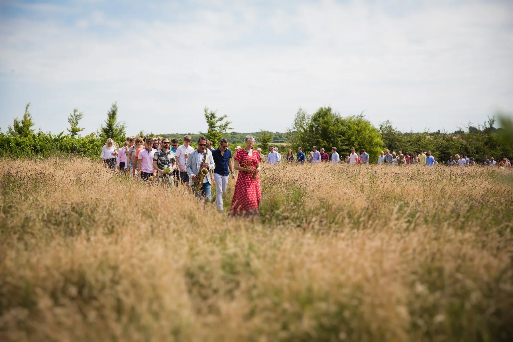 Saxophonist leading a long line of family and friends through the meadow back to the graveside at Clandon Wood natural burial ground in Surrey.