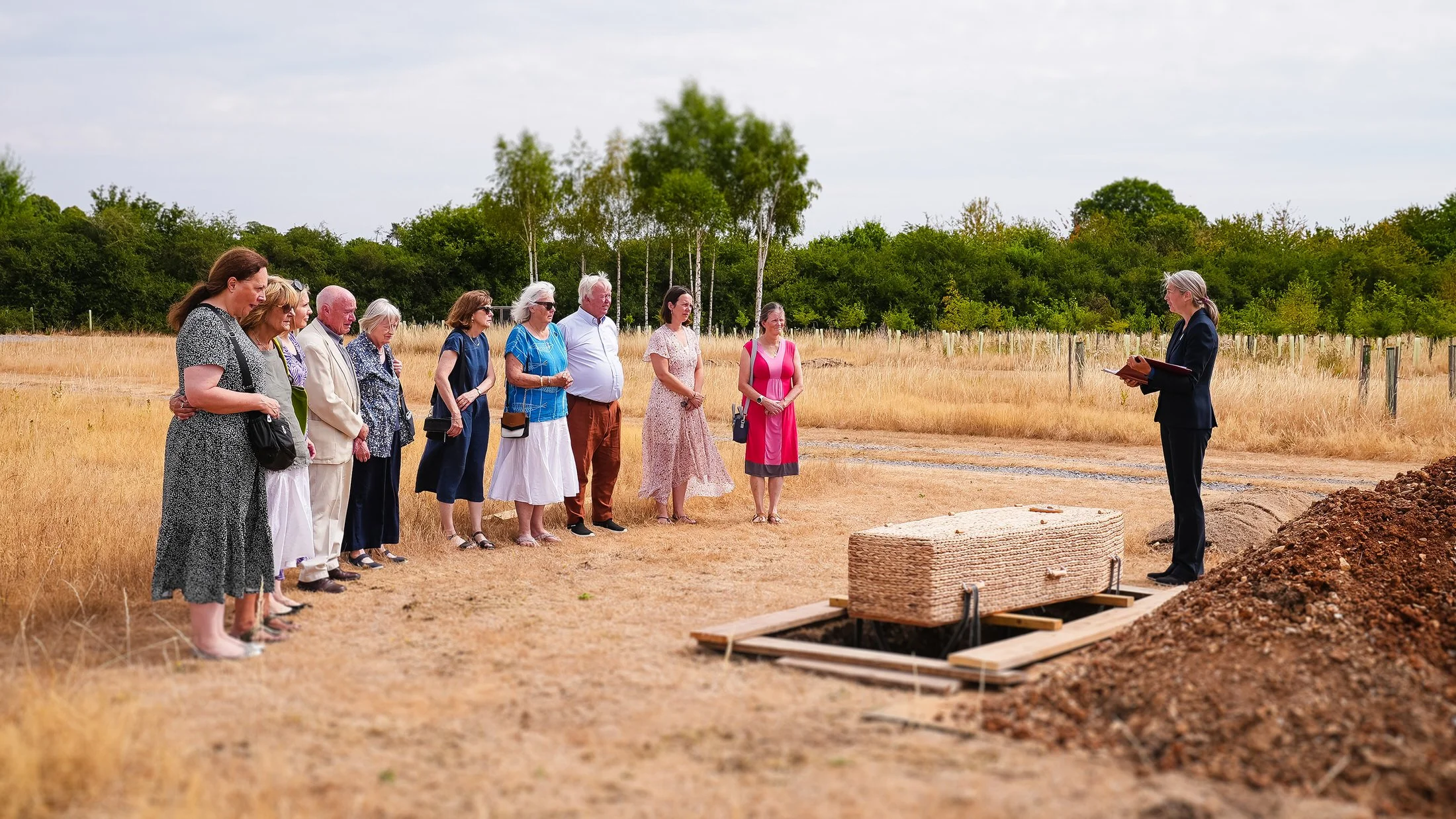 A celebrant leads a natural burial beside a woven coffin as family gather, filmed and photographed by UK Funeral Video Services