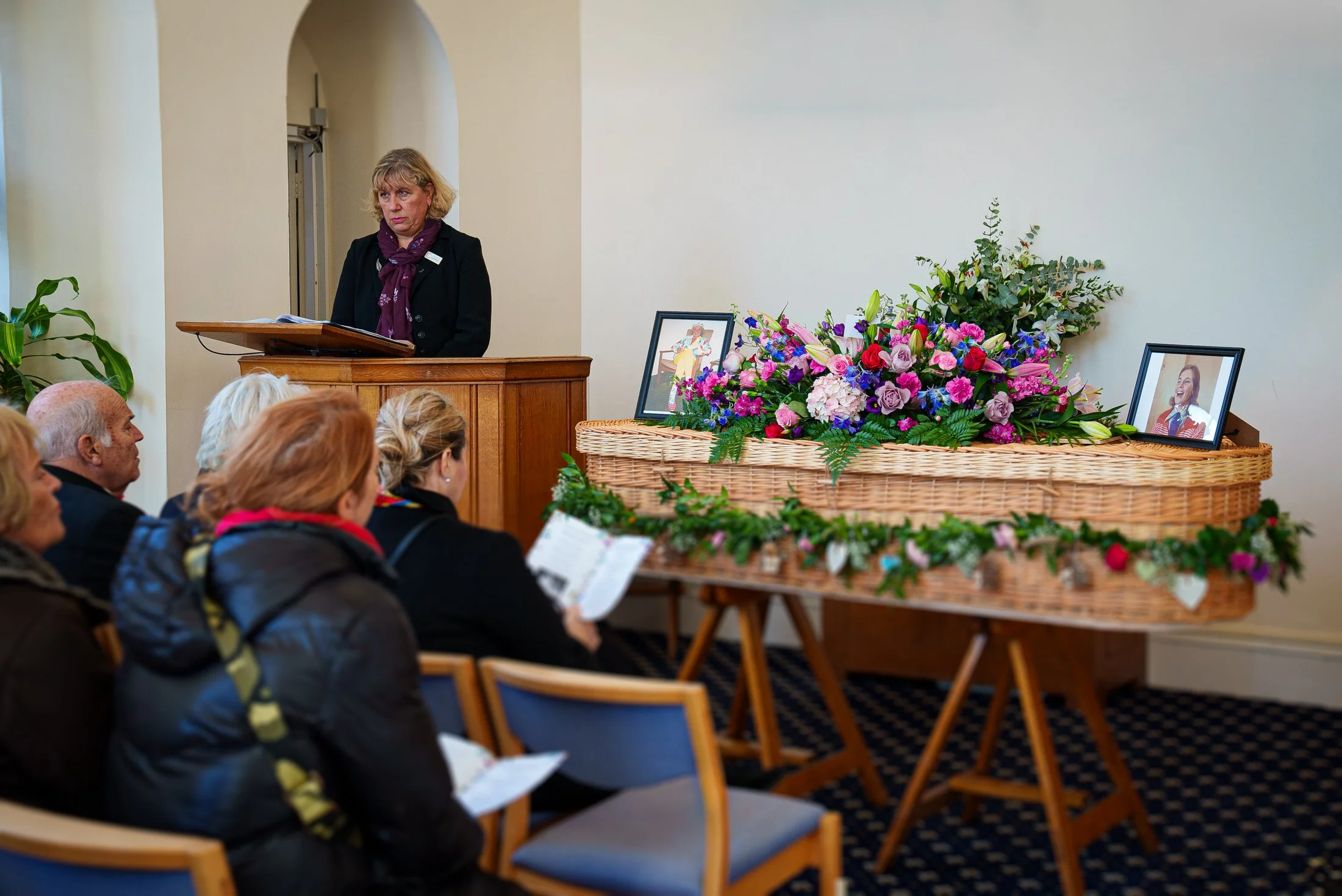 Funeral photography during the service, a celebrant at the lectern beside a flower-covered wicker coffin