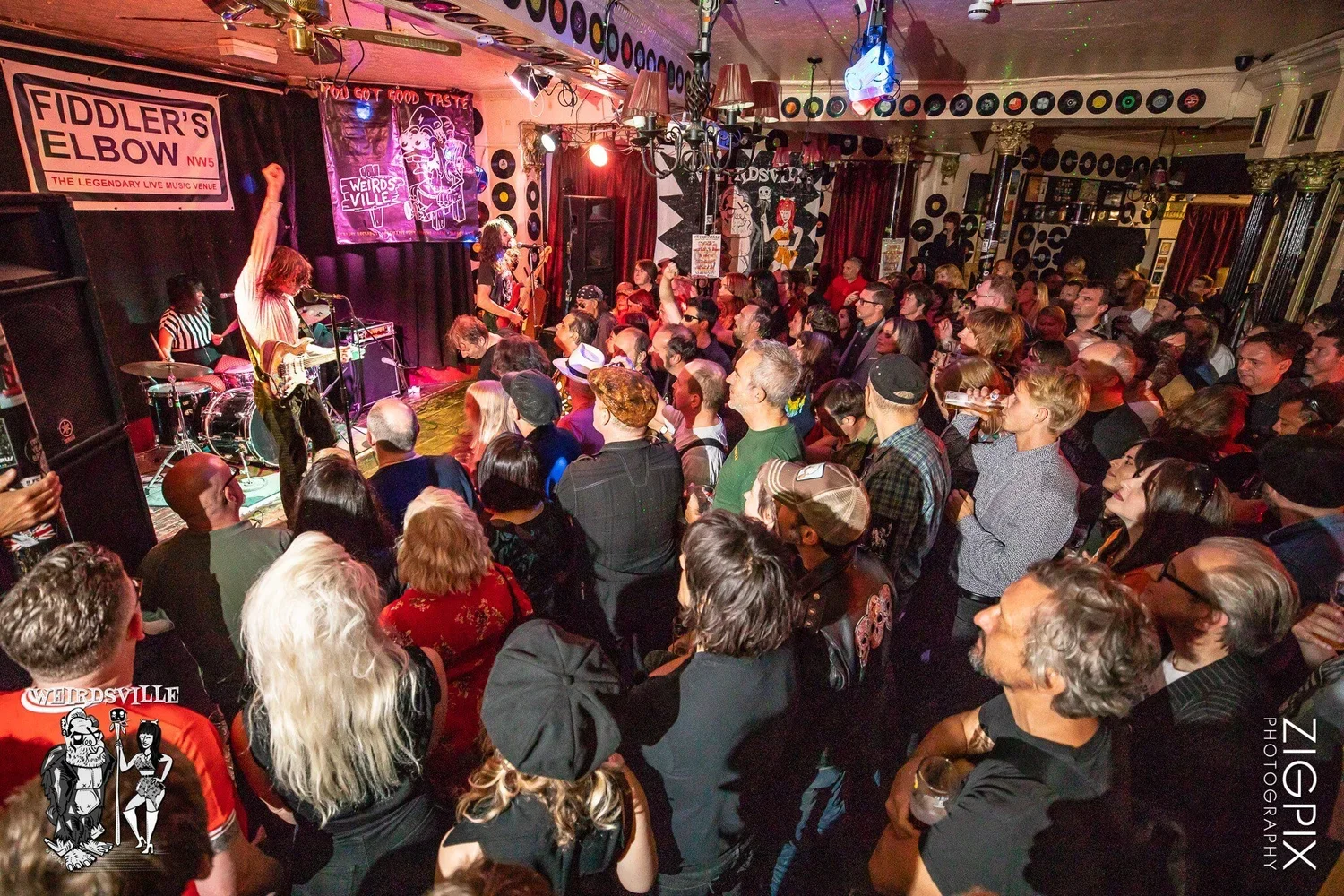 Crowd of people watching live band perform on stage at Fiddler's Elbow music venue decorated with vinyl records and colorful art.