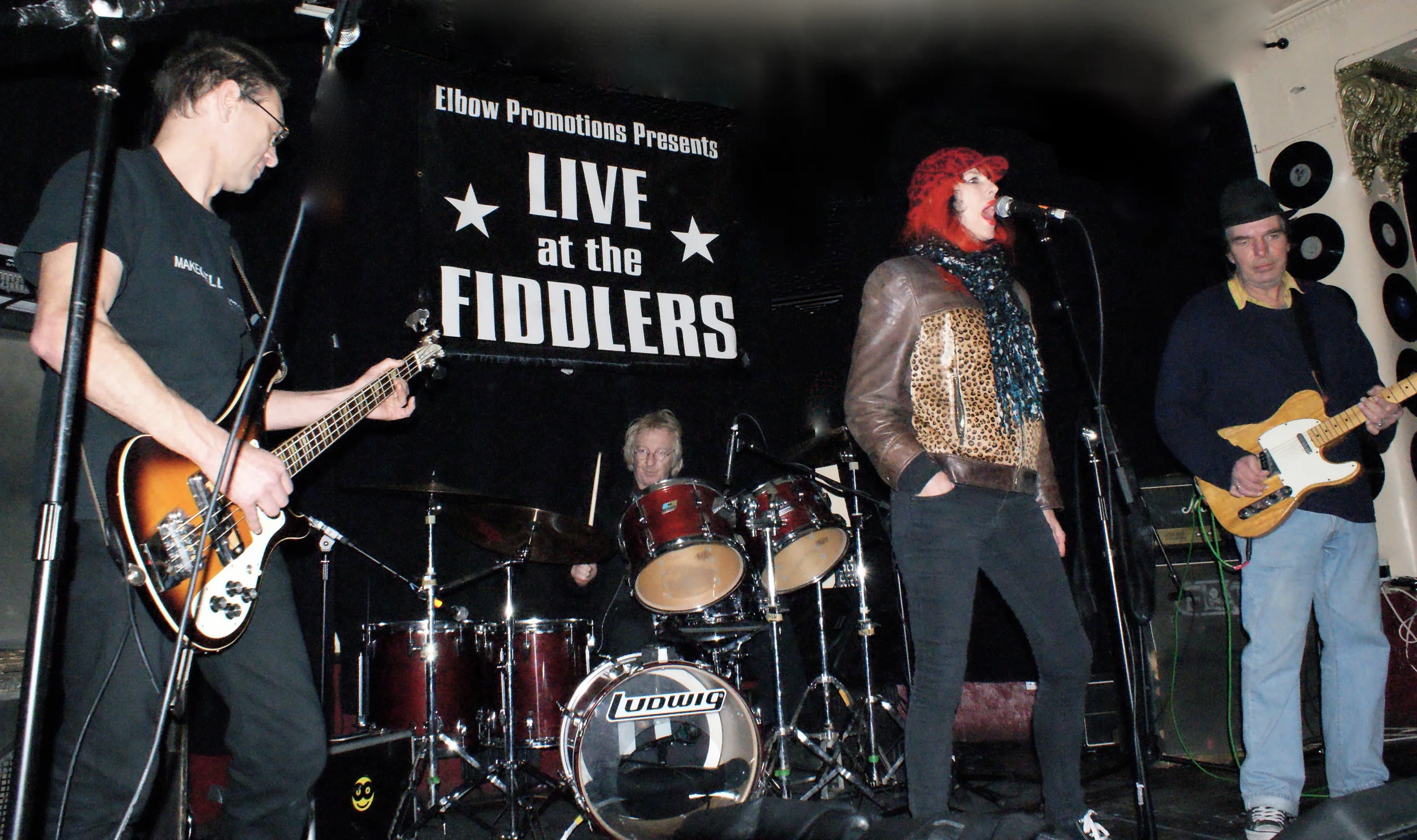 A band performing live on stage at the Fiddlers, with a sign displaying 'Elbow Promotions Presents Live at the Fiddlers' in the background.