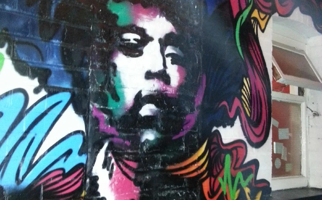 Colorful street art mural of a person's face with highlights in green, purple, and pink, surrounded by vibrant abstract and floral designs.