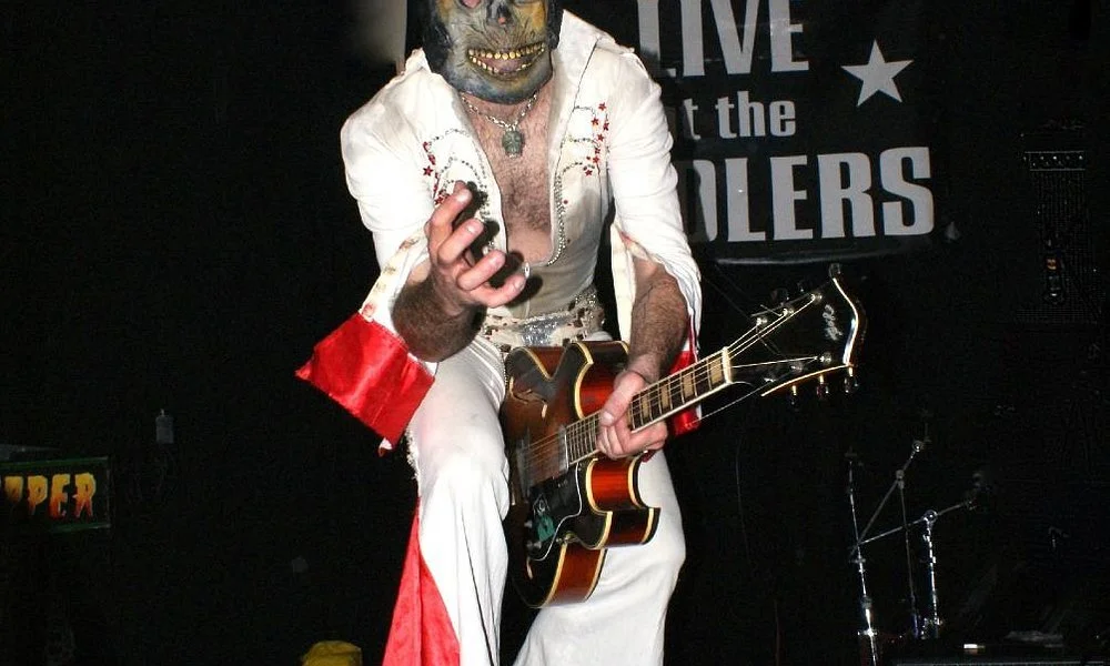 A performer in an Elvis Presley costume holding a guitar and a phone on stage, wearing a mask, with a sign in the background that says 'Live at the Killers'.